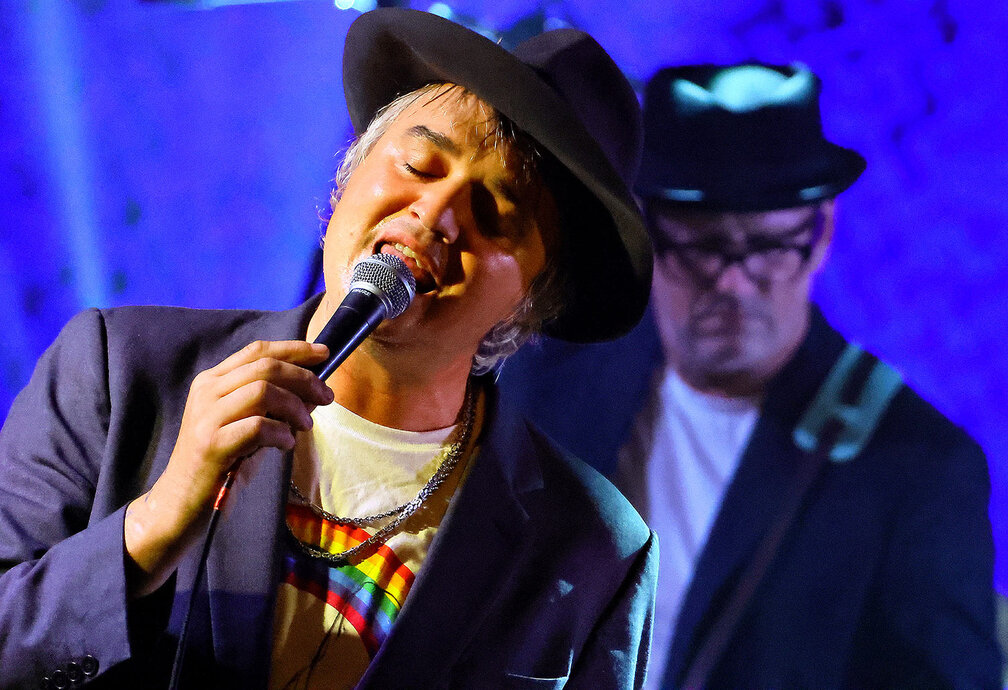 Pete Doherty chantant sur scène avec un microphone sous un éclairage bleu, accompagné d'un musicien en arrière-plan.