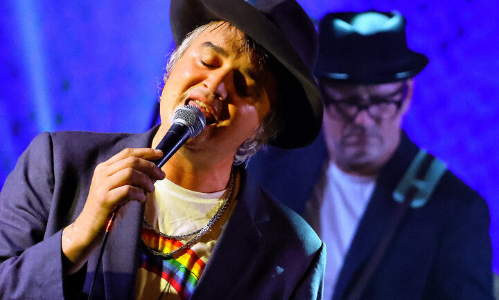 Pete Doherty chantant sur scène avec un microphone sous un éclairage bleu, accompagné d'un musicien en arrière-plan.