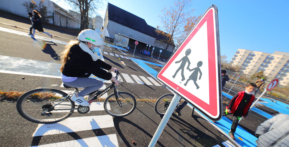 Un groupe d'enfants participe à une activité d'éducation routière dans un espace aménagé en circuit. Au premier plan, une jeune fille portant un casque blanc roule à vélo sur une piste marquée au sol, tandis qu'un panneau triangulaire de signalisation indique un passage fréquent d'enfants. À l'arrière-plan, d'autres enfants, certains à pied et d'autres à vélo, évoluent dans cet environnement pédagogique, sous un ciel dégagé et ensoleillé