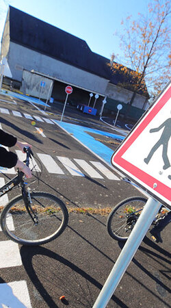 Un groupe d'enfants participe à une activité d'éducation routière dans un espace aménagé en circuit. Au premier plan, une jeune fille portant un casque blanc roule à vélo sur une piste marquée au sol, tandis qu'un panneau triangulaire de signalisation indique un passage fréquent d'enfants. À l'arrière-plan, d'autres enfants, certains à pied et d'autres à vélo, évoluent dans cet environnement pédagogique, sous un ciel dégagé et ensoleillé