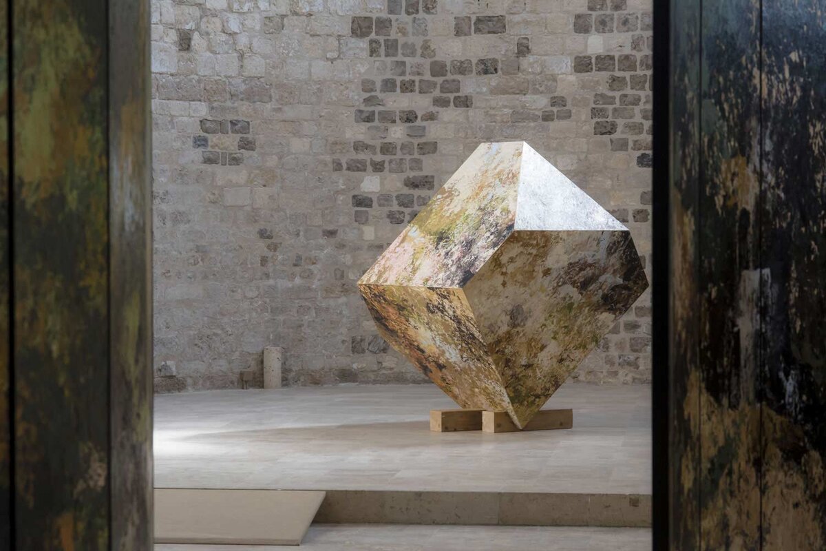 Exposition de Bruno Desplanques à la collégiale Saint-Pierre-le-Puellier d'Orléans. L'image montre une installation artistique contemporaine dans une salle aux murs en pierre apparente. Au centre, une sculpture géométrique abstraite, composée de formes angulaires et de textures minérales, repose sur un socle en bois. L'œuvre est mise en valeur par un éclairage naturel qui accentue ses reliefs et ses nuances. Cette exposition met en lumière le travail unique de Bruno Desplanques, mêlant art moderne et inspiration minérale, dans un cadre historique et intemporel.