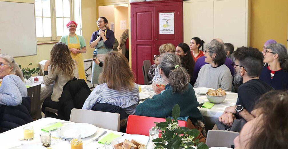 La photo montre un groupe de personnes rassemblées dans une salle lumineuse de la cantine solidaire. Au centre de l'image, deux personnes se tiennent debout, faisant face à l'assemblée. La personne à gauche porte un tablier jaune et un bandana rose, tandis que la personne à droite est vêtue d'un tablier bleu et d'un bandana coloré. Elles semblent s'adresser au groupe, peut-être pour donner des instructions ou partager des informations.  Les participants sont assis autour de tables recouvertes de nappes blanches, sur lesquelles sont disposés des assiettes, des verres et des fleurs. Les personnes présentes semblent attentives et engagées dans la conversation. L'ambiance est conviviale et communautaire, suggérant un espace où les gens se réunissent pour partager un repas et échanger. À l'arrière-plan, une porte rouge est visible, avec une affiche sur le mur à côté.