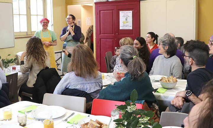 La photo montre un groupe de personnes rassemblées dans une salle lumineuse de la cantine solidaire. Au centre de l'image, deux personnes se tiennent debout, faisant face à l'assemblée. La personne à gauche porte un tablier jaune et un bandana rose, tandis que la personne à droite est vêtue d'un tablier bleu et d'un bandana coloré. Elles semblent s'adresser au groupe, peut-être pour donner des instructions ou partager des informations. Les participants sont assis autour de tables recouvertes de nappes blanches, sur lesquelles sont disposés des assiettes, des verres et des fleurs. Les personnes présentes semblent attentives et engagées dans la conversation. L'ambiance est conviviale et communautaire, suggérant un espace où les gens se réunissent pour partager un repas et échanger. À l'arrière-plan, une porte rouge est visible, avec une affiche sur le mur à côté.