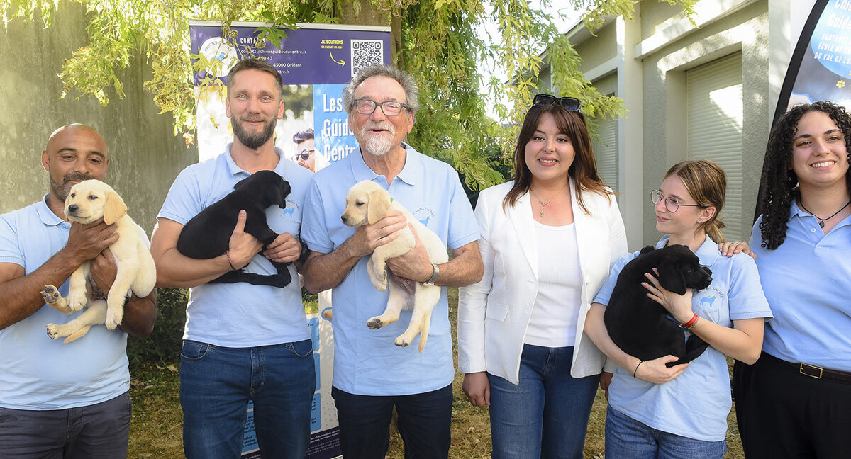 Le président de l'association Michel Brard (au centre) et toute son équipe, accompagnés de l'adjointe au maire, Louna Bailly Photo de groupe en extérieur, dans un jardin ensoleillé. Six personnes posent côte à côte, souriantes, avec quatre chiots labradors, deux beiges et deux noirs. Cinq d'entre elles portent le même polo bleu clair orné d'un petit logo de chien ; la quatrième en partant de la gauche porte une veste blanche, un jean et des lunettes de soleil relevées sur la tête. Au centre, Michel Brard, cheveux gris, lunettes et barbe blanche, tient un chiot beige. À sa gauche, un homme à la barbe courte porte un chiot noir, et un homme crâne rasé, à l'extrémité gauche, tient un chiot beige. À sa droite, la femme en veste blanche a les mains le long du corps, et une jeune femme à lunettes, les cheveux attachés, tient un chiot noir. À l'extrême droite, une femme aux cheveux bouclés sourit à l'objectif. En arrière-plan, une bâche bleue et blanche « Les Chiens guides du Centre » comporte un QR code, et un bâtiment clair se distingue à droite.