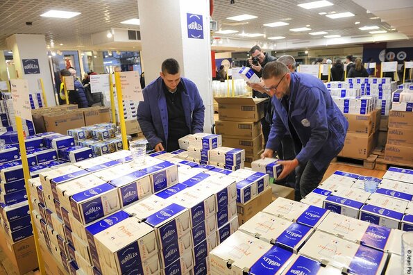 Scène animée à l'intérieur de la boutique Duralex d'Orléans, avec deux employés portant des blouses bleues. Ils manipulent des boîtes empilées de produits Duralex, principalement des verres en carton d'emballage bleu et blanc