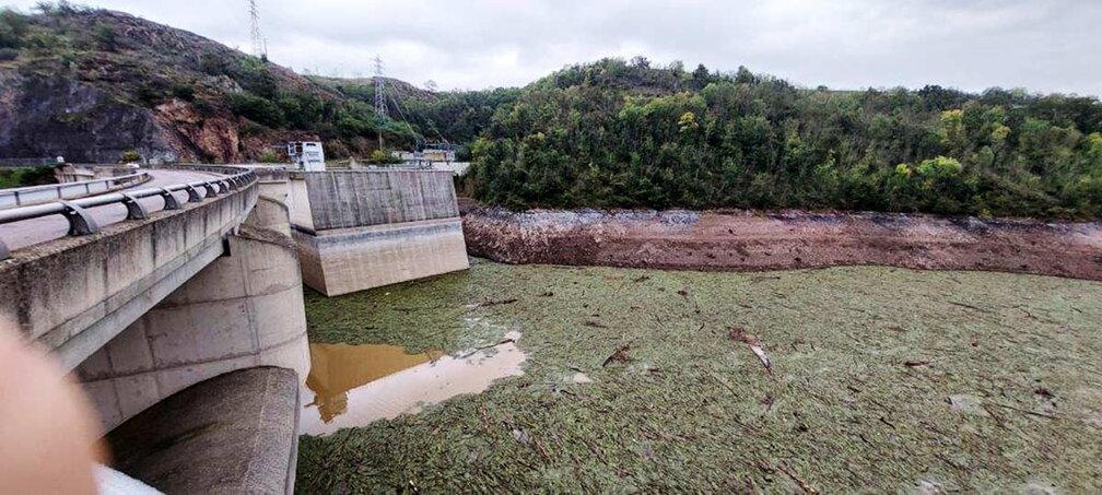 Vue d'un barrage en béton avec une structure en arc à gauche, situé dans un environnement naturel avec une colline boisée en arrière-plan. À l'avant-plan, une grande étendue d'eau recouverte de débris flottants, principalement des végétaux et des branches, après une crue de la Loire. Le ciel est nuageux, et des pylônes électriques sont visibles sur la colline
