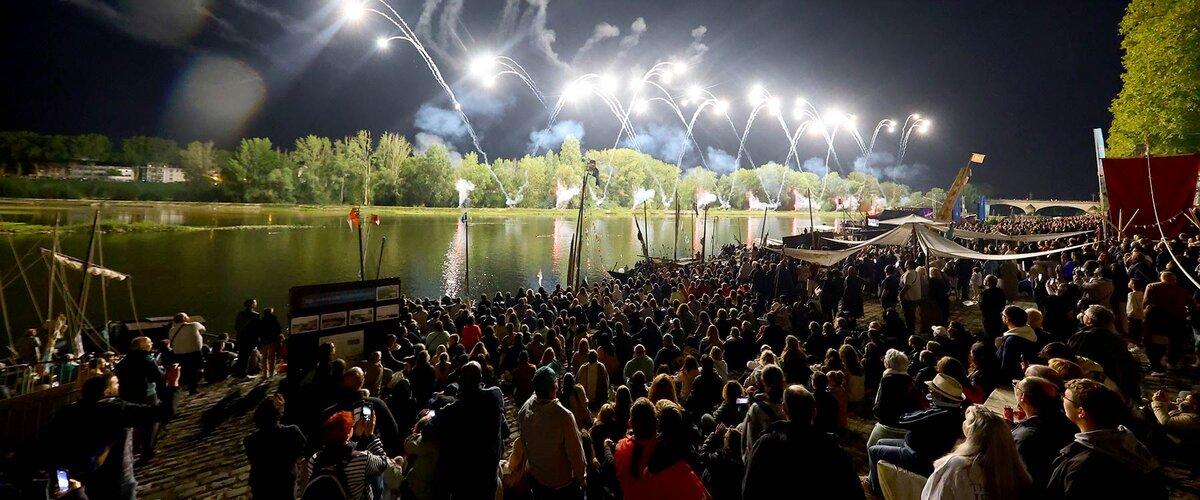 Vue nocturne du spectacle pyro-symphonique sur la Loire pendant le Festival de Loire. La foule dense est rassemblée sur les berges, regardant des feux d’artifice s’élever dans le ciel au-dessus du fleuve, éclairant la scène et les arbres en arrière-plan. Des embarcations traditionnelles et des installations de type médiéval bordent la rive, créant une ambiance festive et immersive.