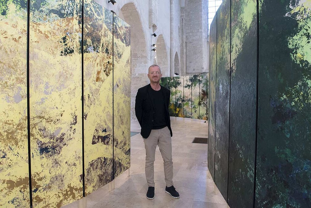 Sur cette photo, Bruno Desplanques, vêtu d’une tenue sobre composée d’un blazer noir, d’un t-shirt assorti et d’un pantalon beige, se tient debout au milieu d’une installation artistique immersive. L’espace, baigné de lumière naturelle, est une vaste salle aux hautes voûtes en pierre, évoquant l’architecture d’un édifice historique.  Autour de lui, de grands panneaux peints, aux couleurs et textures abstraites, créent une atmosphère entre nature et abstraction. Les tons oscillent entre des nuances de vert profond, de jaune doré et des touches plus sombres, conférant à l’ensemble une impression de paysage fragmenté. L’installation semble transformer l’espace en une œuvre vivante, où les visiteurs peuvent circuler et se laisser envelopper par les jeux de couleurs et de matières.  Le lieu, la collégiale Saint-Pierre-le-Puellier d’Orléans, offre un contraste saisissant entre l’art contemporain et la pierre ancienne, soulignant la rencontre entre patrimoine et création artistique moderne.