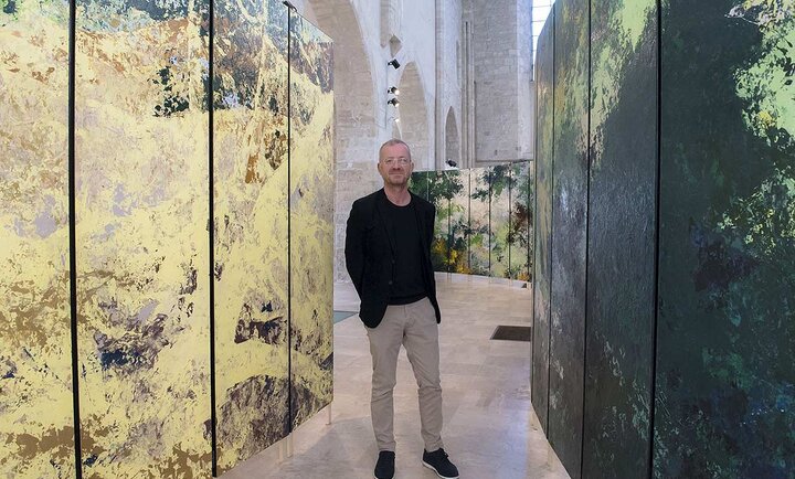 Sur cette photo, Bruno Desplanques, vêtu d’une tenue sobre composée d’un blazer noir, d’un t-shirt assorti et d’un pantalon beige, se tient debout au milieu d’une installation artistique immersive. L’espace, baigné de lumière naturelle, est une vaste salle aux hautes voûtes en pierre, évoquant l’architecture d’un édifice historique. Autour de lui, de grands panneaux peints, aux couleurs et textures abstraites, créent une atmosphère entre nature et abstraction. Les tons oscillent entre des nuances de vert profond, de jaune doré et des touches plus sombres, conférant à l’ensemble une impression de paysage fragmenté. L’installation semble transformer l’espace en une œuvre vivante, où les visiteurs peuvent circuler et se laisser envelopper par les jeux de couleurs et de matières. Le lieu, la collégiale Saint-Pierre-le-Puellier d’Orléans, offre un contraste saisissant entre l’art contemporain et la pierre ancienne, soulignant la rencontre entre patrimoine et création artistique moderne.