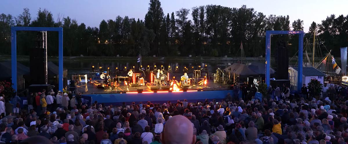 scène de concert en soirée au Festival de Loire en 2023, avec une foule nombreuse rassemblée devant la scène du ponton sur les quais de Loire. La scène est bien éclairée avec des lumières colorées mettant en valeur les musiciens et leurs instruments. L'ambiance semble festive et animée, avec des spectateurs attentifs profitant du spectacle de François Morel.