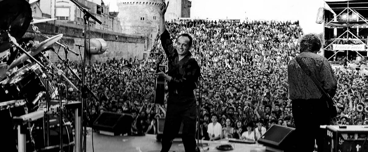 Photo en noir et blanc du groupe Au Petit Bonheur en concert sur la Grande Scène de Saint Jean d'Acre à La Rochelle, devant une immense foule enthousiaste. Le chanteur principal, souriant et levant un bras avec sa guitare, salue le public. À ses côtés, un autre membre du groupe joue également de la guitare. À gauche, on aperçoit une batterie. L’ambiance est festive et l’arrière-plan révèle les remparts historiques de la ville.