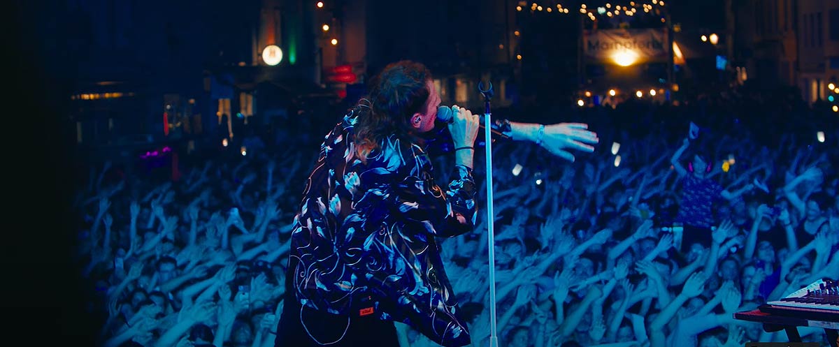 Le chanteur Valentino Vivace est en train de se produire sur scène lors d'un concert nocturne. Il est vêtu d'une chemise à motifs sombres et se tient près d'un micro, captivant le public avec sa performance. La foule, visible en contrebas, lève les mains en signe d'enthousiasme, et les lumières de la scène éclairent l'atmosphère énergique du spectacle.