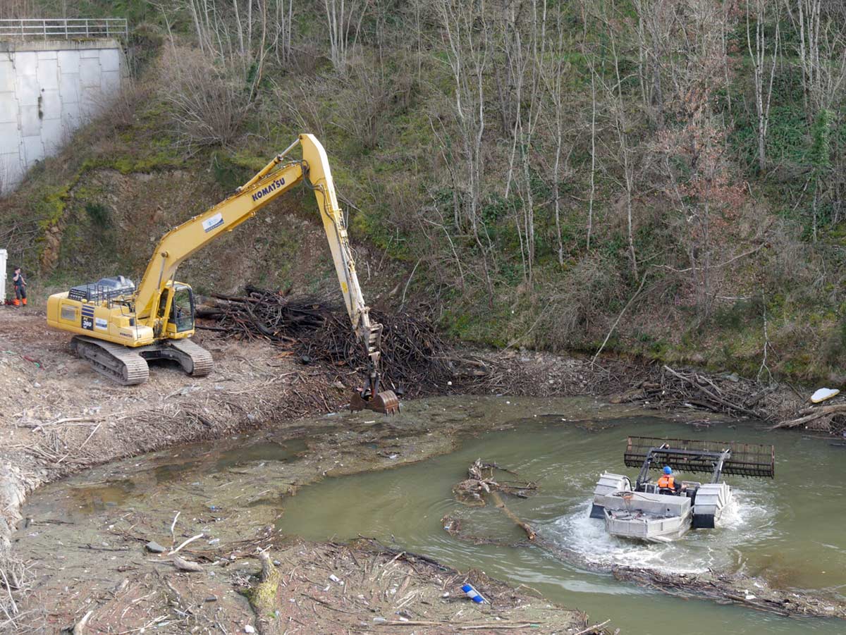 Une pelle mécanique jaune Komatsu est positionnée sur une berge en pente, près d'un cours d'eau. Elle est en train de déplacer des branches et des débris végétaux. En arrière-plan, une forêt d'arbres sans feuilles est visible. Sur le cours d'eau, une petite embarcation motorisée avec deux personnes à bord navigue près de la rive. La scène se déroule au barrage de Villerest pendant une opération de nettoyage.