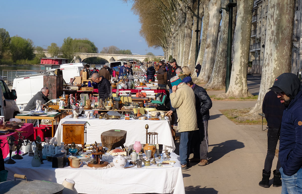Une brocante animée se tient sur le quai du Châtelet à Orléans par une belle journée ensoleillée. De longues rangées de stands débordent d’objets anciens et de curiosités : vaisselle, lampes, livres, meubles et bibelots variés. Des chineurs, vêtus chaudement, fouillent avec attention parmi les trésors exposés, échangeant avec les vendeurs. En arrière-plan, les platanes alignés longent le quai, et le pont sur la Loire ajoute une touche pittoresque au décor. L’ambiance est conviviale et propice aux découvertes.